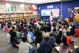  A lady stands in a library talking to a large group of young children sitting down.